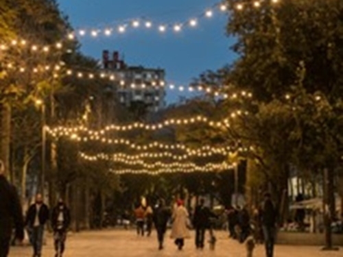 Photograph of a brick street, lined with trees, people walking, and string lights in a zig-zag pattern.