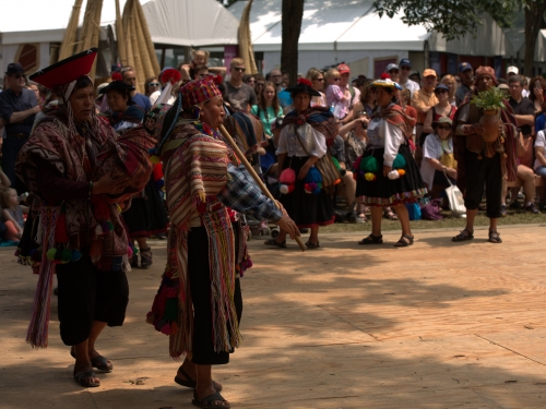A man in a vibrant colorful outfit plays a long wooden flute in front of a crowd.