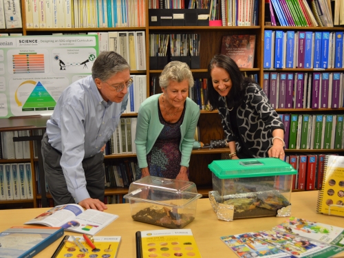 Two women and a man stand in a classroom, leaning over some desks with clear animal crates on them.